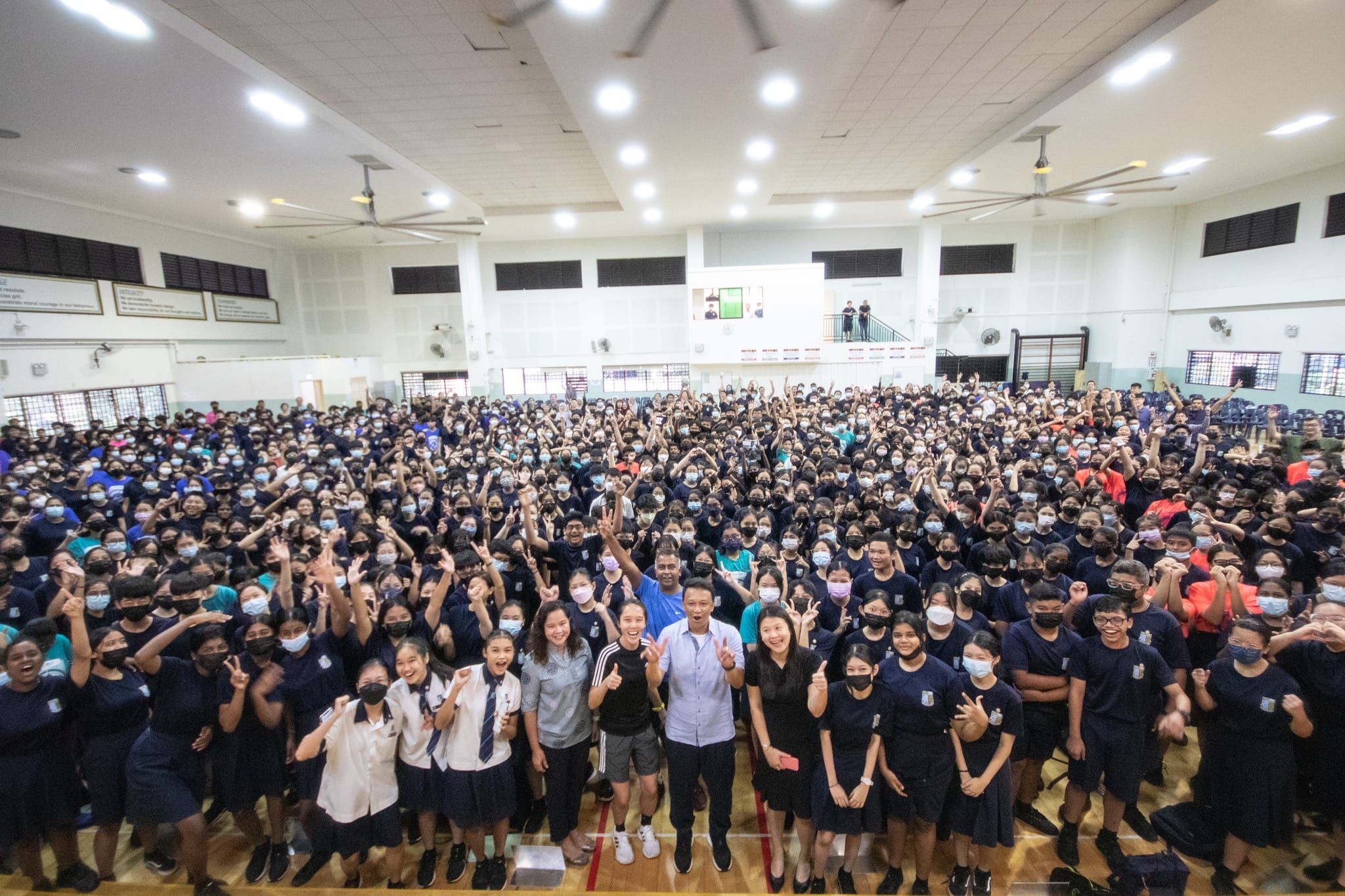 A large crowd of school students in navy uniforms wearing masks fill an indoor hall, cheering and posing for a group photo.